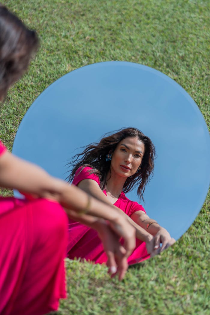 A woman in a red dress gazes into a mirror outdoors, capturing elegance and reflection.